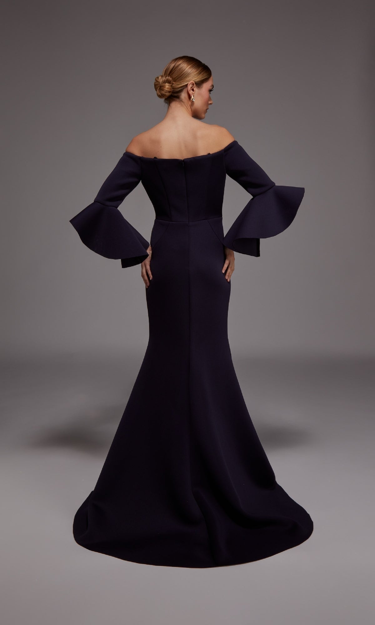 A woman stands with her back to the camera in a dark, off-the-shoulder Long Formal Dress: Alyce 27777 featuring flared sleeves, set against a plain gray background.